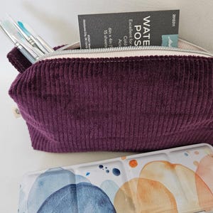 May include: A deep purple corduroy zippered pouch containing several paintbrushes and a small pad of watercolor paper. In front of the pouch sits a rectangular metal watercolor paint tin featuring an abstract blue and orange design.