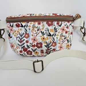 May include: A floral print fanny pack with a white strap and bronze-colored hardware. The bag features a brown zipper and is covered in a pattern of flowers in shades of red, pink, yellow, and green on a white background.