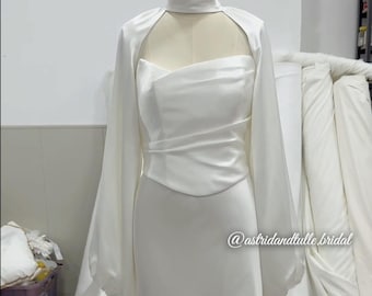 Puede incluir: Vestido de novia de satén blanco sobre un maniquí. El vestido presenta un cuello alto, mangas largas y un corpiño ajustado que fluye hacia una falda larga con cola. El vestido tiene un top estilo bolero. El texto "@astridandtulle.bridal" es visible.