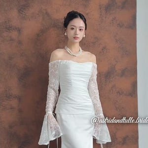 May include: A white off-the-shoulder wedding dress with long lace sleeves and a flowing skirt. The dress features a fitted bodice and a subtle train. The background is a brown wall. The dress is elegant and suitable for a wedding.