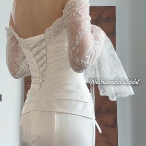 May include: A white wedding dress with a corset back and off-the-shoulder lace sleeves. The dress has a fitted bodice and a long skirt. The sleeves are long and made of delicate lace. The dress is elegant and suitable for a wedding.