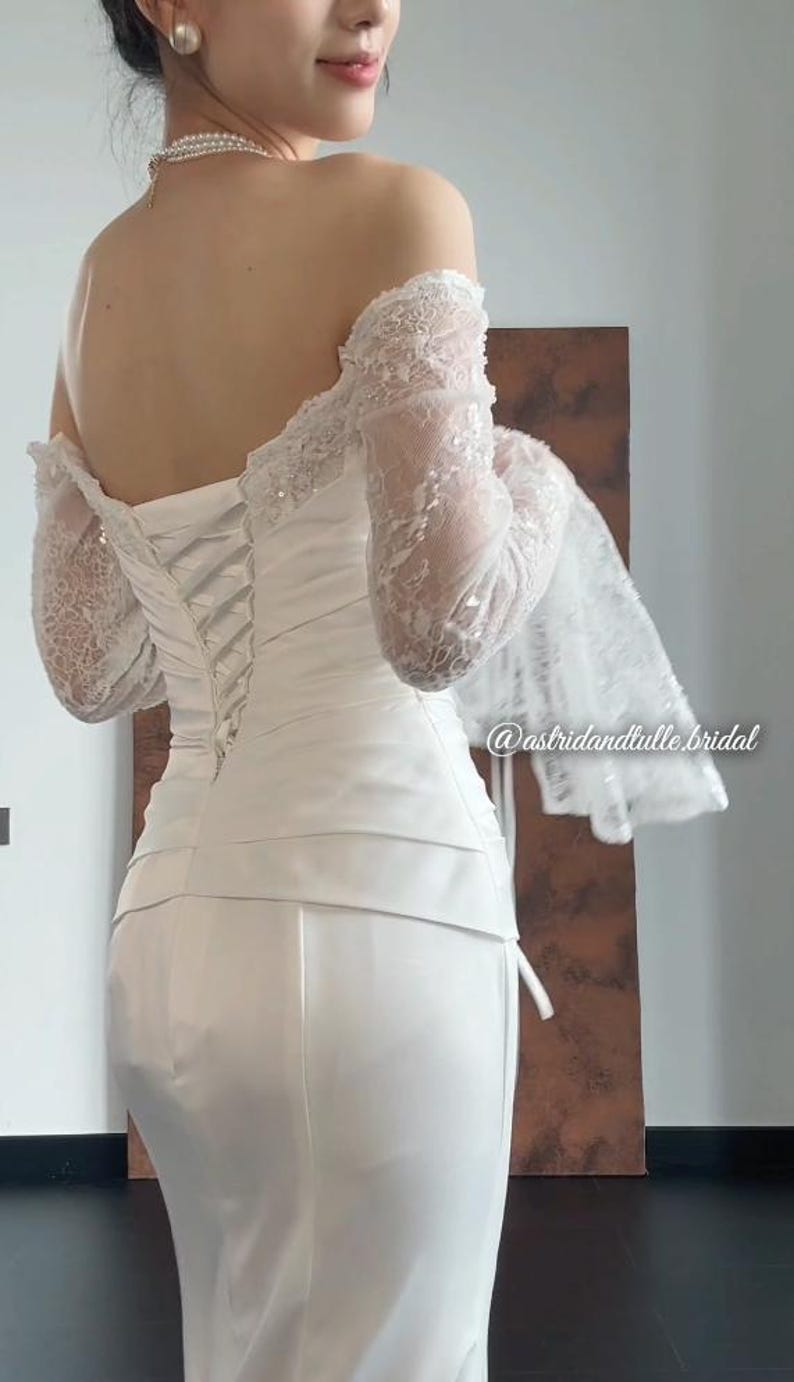 May include: A white wedding dress with a corset back and off-the-shoulder lace sleeves. The dress has a fitted bodice and a long skirt. The sleeves are long and made of delicate lace. The dress is elegant and suitable for a wedding.