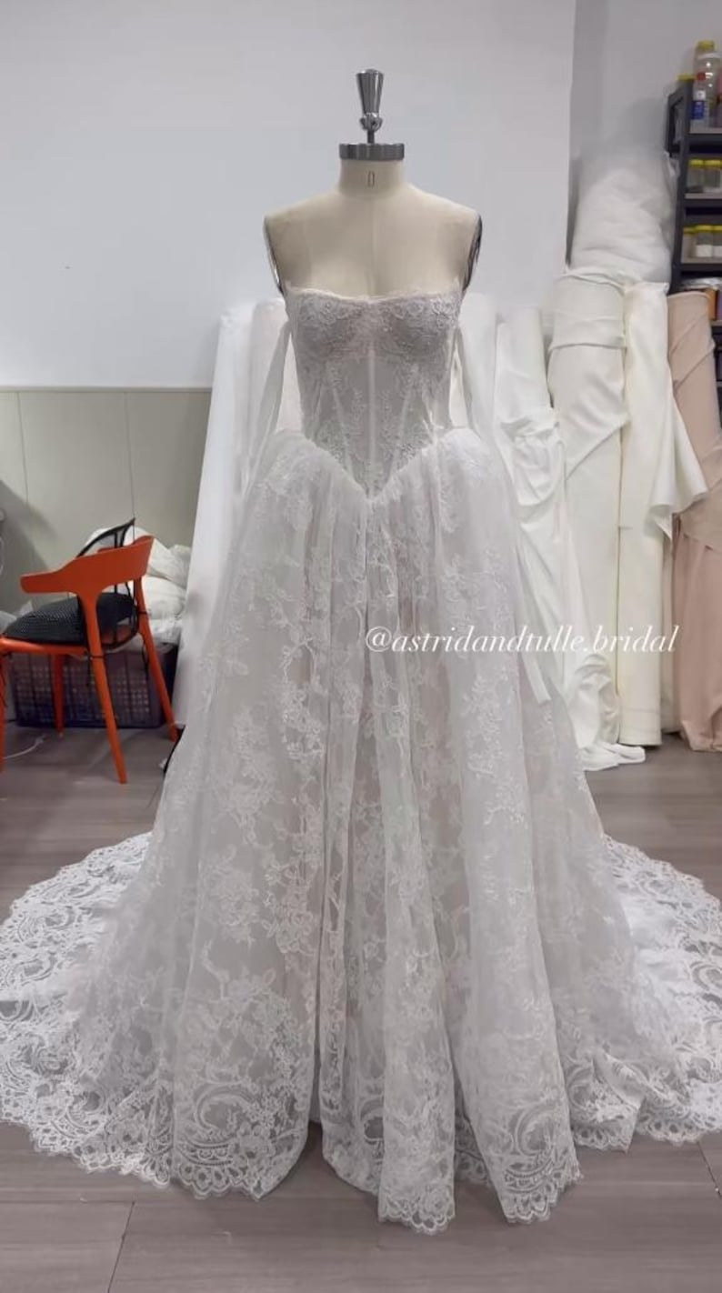 May include: A white strapless wedding dress on a mannequin featuring a corset-style bodice with intricate lace overlay and a full, floor-length skirt with a long train. The text @astridandtulle.bridal is visible on the front of the gown.