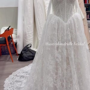 May include: A white strapless wedding dress on a mannequin featuring a corset-style bodice with intricate floral lace detailing, a full skirt with a long lace-trimmed train, and the text @astridandtulle.bridal printed on the front of the skirt.