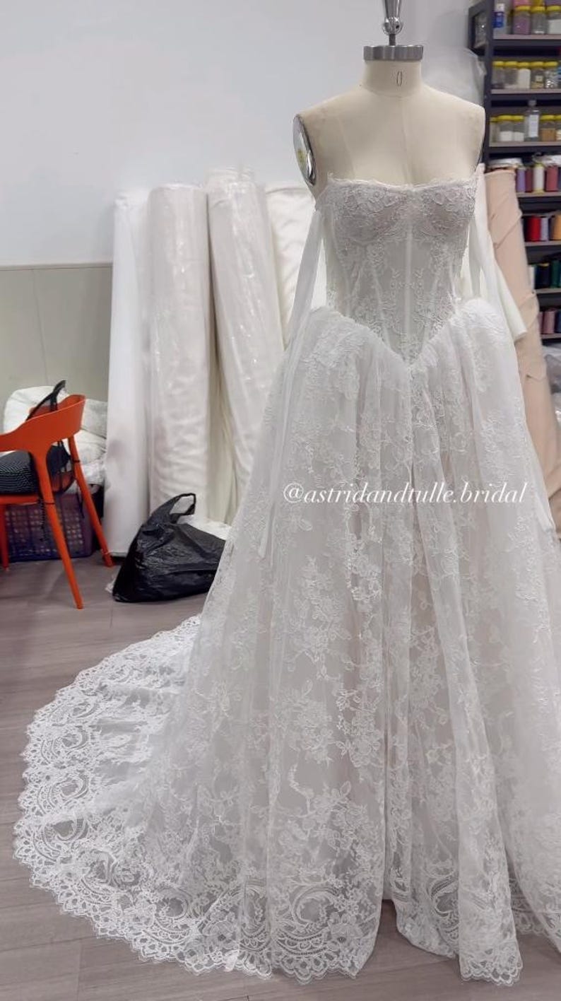 May include: A white strapless wedding dress on a mannequin featuring a corset-style bodice with intricate floral lace detailing, a full skirt with a long lace-trimmed train, and the text @astridandtulle.bridal printed on the front of the skirt.