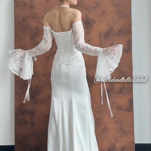 May include: A white bridal gown with a fitted bodice and a flowing skirt. The dress features off-the-shoulder sleeves with lace detailing and bell-shaped cuffs. The back of the dress has a corset-style closure. The image includes the text "@astridandtulle.bridal".
