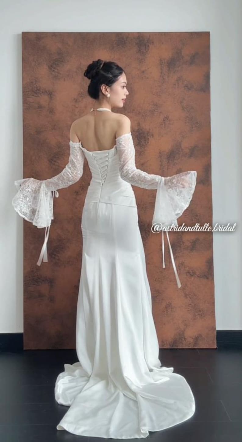 May include: A white bridal gown with a fitted bodice and a flowing skirt. The dress features off-the-shoulder sleeves with lace detailing and bell-shaped cuffs. The back of the dress has a corset-style closure. The image includes the text "@astridandtulle.bridal".