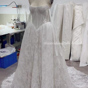 May include: A white strapless floor-length gown featuring a structured corset bodice with intricate lace detailing and a full skirt with a long train. The dress displays the text @astridandtulle.bridal across the midsection of the skirt.