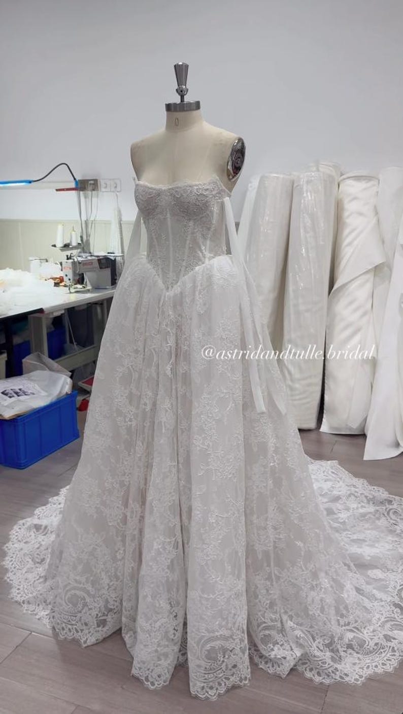 May include: A white strapless floor-length gown featuring a structured corset bodice with intricate lace detailing and a full skirt with a long train. The dress displays the text @astridandtulle.bridal across the midsection of the skirt.