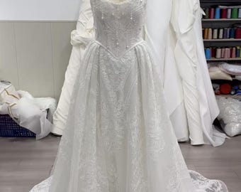 Op de afbeelding: Een witte trouwjurk op een mannequin. De strapless lijfje is versierd met delicate kralen en kant. De rok heeft ingewikkelde kanten details en een lange sleep. De jurk wordt getoond in een studio met andere witte stoffen op de achtergrond.