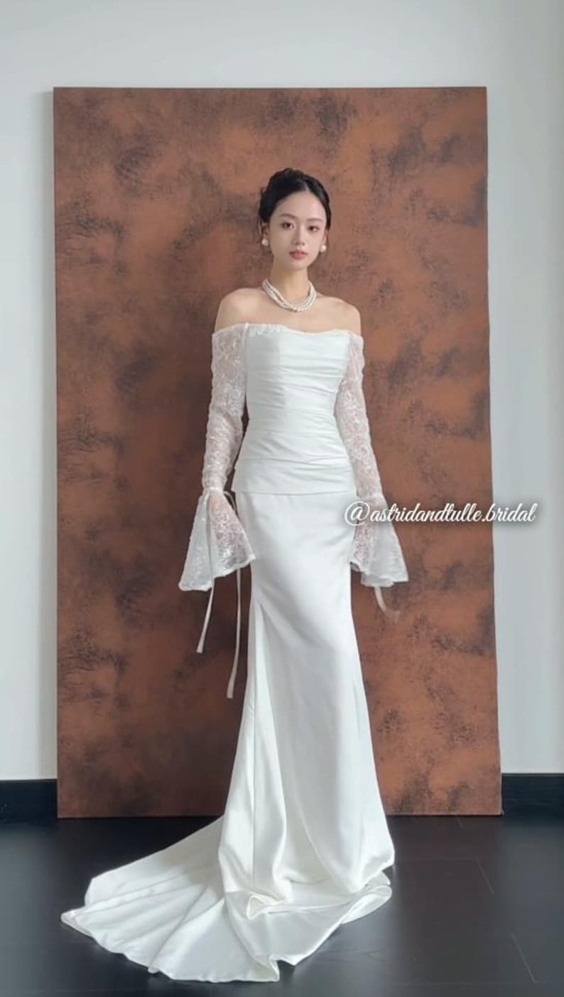 May include: A white off-the-shoulder wedding dress with long lace sleeves and a flowing skirt. The dress features a fitted bodice and a subtle train. The background is a brown wall. The dress is elegant and suitable for a wedding.