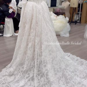 May include: A floor-length white lace wedding dress on a mannequin, featuring an off-the-shoulder neckline, long sleeves, a corset-style lace-up back, and a full skirt with a long train. The text @astridandtulle.bridal is visible on the image.