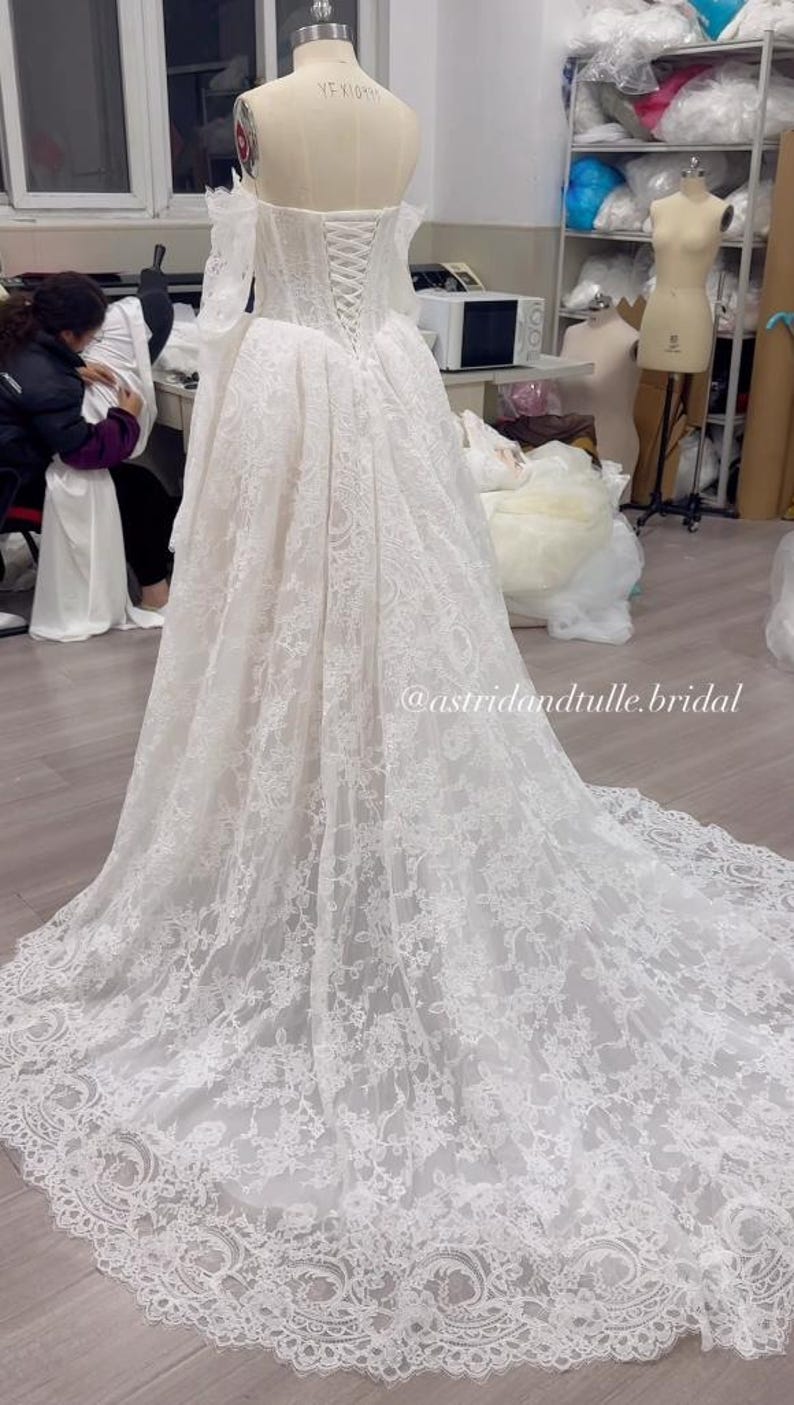 May include: A floor-length white lace wedding dress on a mannequin, featuring an off-the-shoulder neckline, long sleeves, a corset-style lace-up back, and a full skirt with a long train. The text @astridandtulle.bridal is visible on the image.