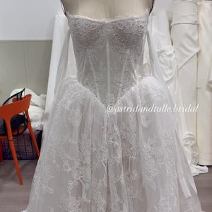 May include: A white strapless gown on a mannequin featuring a corset-style bodice with intricate floral lace overlays, a sweetheart neckline, and a full skirt with matching lace detailing. The text @astridandtulle.bridal is visible.