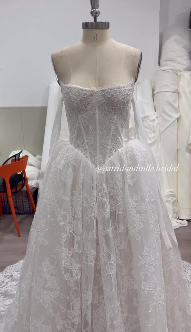 May include: A white strapless gown on a mannequin featuring a corset-style bodice with intricate floral lace overlays, a sweetheart neckline, and a full skirt with matching lace detailing. The text @astridandtulle.bridal is visible.