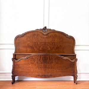 May include: A wooden bed frame featuring a tall, arched headboard and a lower footboard, both crafted from rich brown wood with prominent burl grain patterns. The frame is adorned with intricate carved scrollwork details along the top edges.