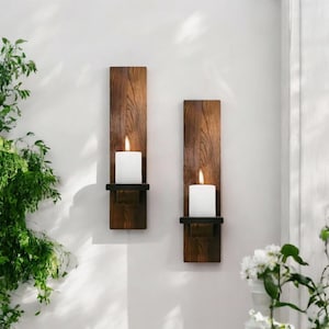 May include: Two wooden wall sconces with a dark brown finish, each holding a white candle with a lit flame. The sconces are mounted on a white wall, with greenery and white flowers in the background.