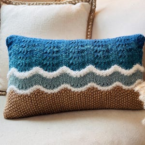 May include: A knitted throw pillow with a blue, white, and brown wave pattern. The pillow is on a white couch.