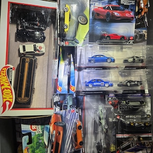 May include: A collection of various Hot Wheels die-cast toy cars in their original blister packaging and a boxed set. The assortment includes multiple sports cars, sedans, and racing vehicles in diverse colors like red, blue, orange, and black.