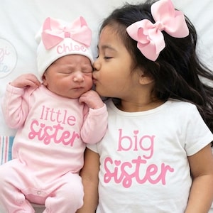 May include: A light pink long-sleeve onesie with the text little sister in pink, a white beanie with a pink bow and the name Chloe, and a white short-sleeve t-shirt with the text big sister in pink and a large pink hair bow.