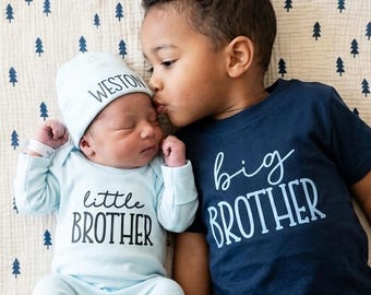 May include: A newborn baby wearing a light blue onesie and cap with the text "little BROTHER" and "WESTON" respectively, lying next to a child in a navy blue shirt with "big BROTHER" printed on it. The children are on a cream-colored blanket with a blue tree pattern.