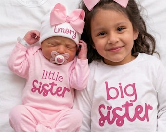 May include: A baby wearing a pink long-sleeve onesie with the text little sister and a white hat with the name Emory and a pink bow, next to a child wearing a white long-sleeve shirt with the text big sister and a pink hair bow.