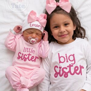 May include: A baby wearing a pink long-sleeve onesie with the text little sister and a white hat with the name Emory and a pink bow, next to a child wearing a white long-sleeve shirt with the text big sister and a pink hair bow.