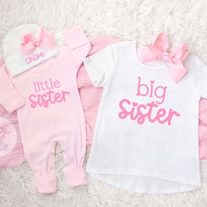 May include: A light pink long-sleeve romper with the text little sister, a white beanie with a pink bow and the name Chloe, and a white short-sleeve t-shirt with the text big sister and a pink bow. The text is in a pink script font.
