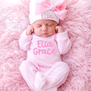 May include: A white newborn hat with a large pink bow and the name Elia embroidered in pink, paired with a light pink long-sleeved footed sleeper featuring the name Elia Grace printed in a bold pink script font across the chest area.