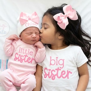 May include: A light pink long-sleeve onesie with the text little sister in pink, a white beanie with a pink bow and the name Chloe, and a white short-sleeve t-shirt with the text big sister in pink and a large pink hair bow.