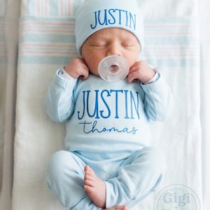May include: A newborn baby wearing a light blue hat and onesie, both with the name "JUSTIN" printed in blue. The baby is also wearing a pacifier and is lying on a white and blue striped blanket.