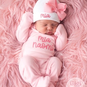 May include: A light pink long-sleeved footed sleeper with the name Mia Naomi printed in pink on the chest. A white beanie with a large pink bow and the name Mia embroidered in pink is worn on the head. The outfit is made of soft fabric.