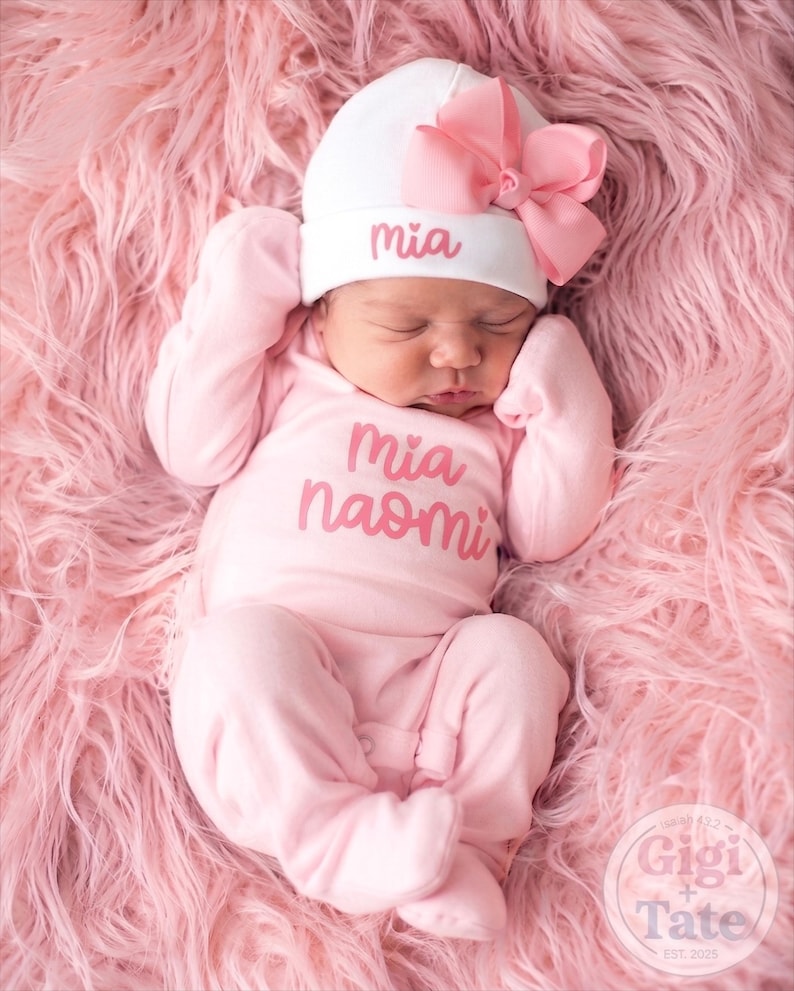 May include: A light pink long-sleeved footed sleeper with the name Mia Naomi printed in pink on the chest. A white beanie with a large pink bow and the name Mia embroidered in pink is worn on the head. The outfit is made of soft fabric.