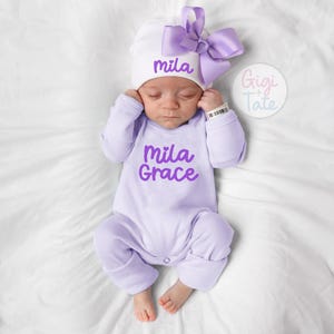 May include: A light purple long-sleeve infant bodysuit with the name Mila Grace printed in purple on the chest. The infant wears a white beanie featuring a large purple bow and the name Mila printed in purple on the front of the hat.