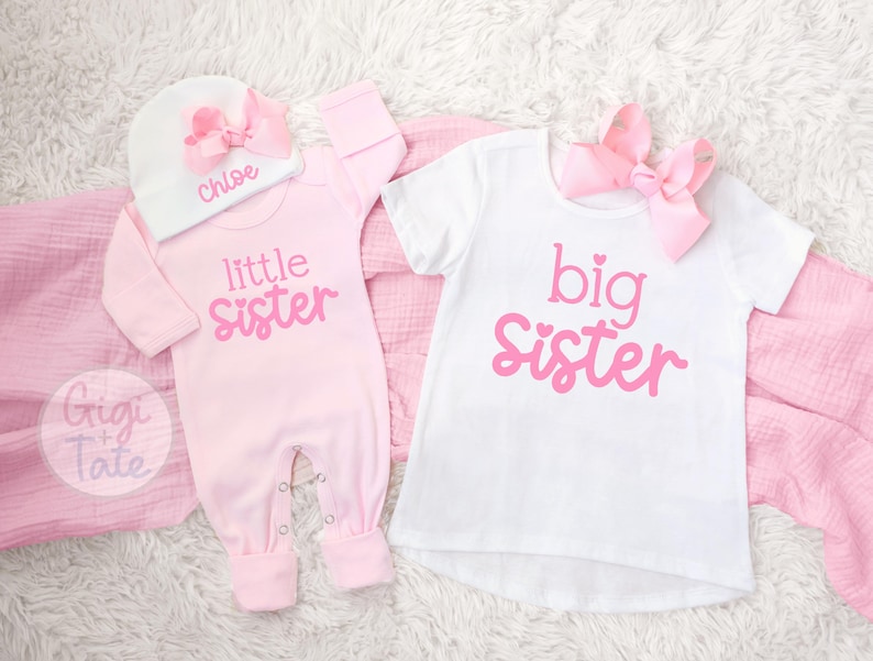 May include: A light pink long-sleeve romper with the text little sister, a white beanie with a pink bow and the name Chloe, and a white short-sleeve t-shirt with the text big sister and a pink bow. The text is in a pink script font.