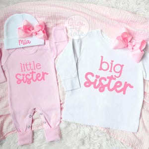 May include: A light pink long-sleeve onesie with the text little sister in pink, paired with a white beanie featuring a pink bow and the name Mia. Beside it, a white long-sleeve shirt with the text big sister in pink and a pink bow.