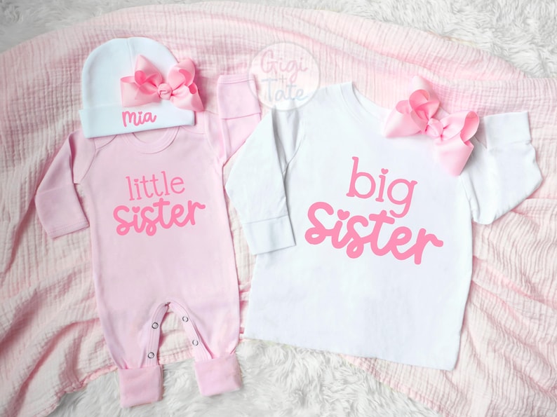 May include: A light pink long-sleeve onesie with the text little sister in pink, paired with a white beanie featuring a pink bow and the name Mia. Beside it, a white long-sleeve shirt with the text big sister in pink and a pink bow.