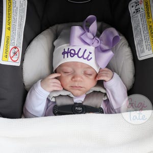 May include: A white baby hat featuring the name Holli embroidered in purple script with a small heart above the letter i. A large, lavender-colored ribbon bow is attached to the side of the hat. The hat is worn by a sleeping infant in a car seat.