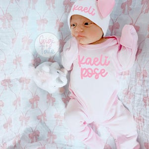 May include: A white baby onesie and matching hat with a large pink bow, both personalized with the name Kaeli Rose in pink script. The onesie features long sleeves and footed bottoms. A small white plush unicorn toy is positioned nearby.
