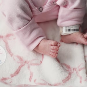 May include: A soft white baby blanket featuring a repeating pattern of light pink bows. The blanket is textured and plush. A circular logo with the text Gigi + Tate is visible on the left side. A baby wearing a light pink long-sleeve garment rests on the blanket, with a small white hospital identification band visible on the ankle.