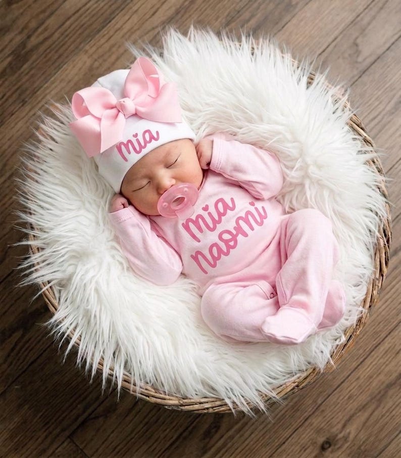 May include: A white baby hat with a large pink bow and the name Mia embroidered in pink, paired with a light pink long-sleeved onesie featuring the name Mia Naomi in pink script, and a clear pink pacifier worn by a sleeping infant.