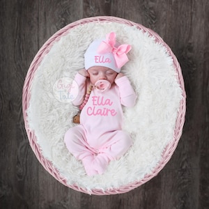 May include: A newborn baby wearing a light pink long-sleeve footed sleeper and a white beanie with a large pink bow. The beanie features the name Ella in pink script, and the sleeper displays the name Ella Claire in pink script with a small heart.
