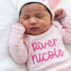 May include: A white newborn hat featuring a light pink bow and the name River embroidered in pink, paired with a long-sleeved light pink onesie printed with the name River Nicole in a bold pink script font with small heart accents over the i letters.