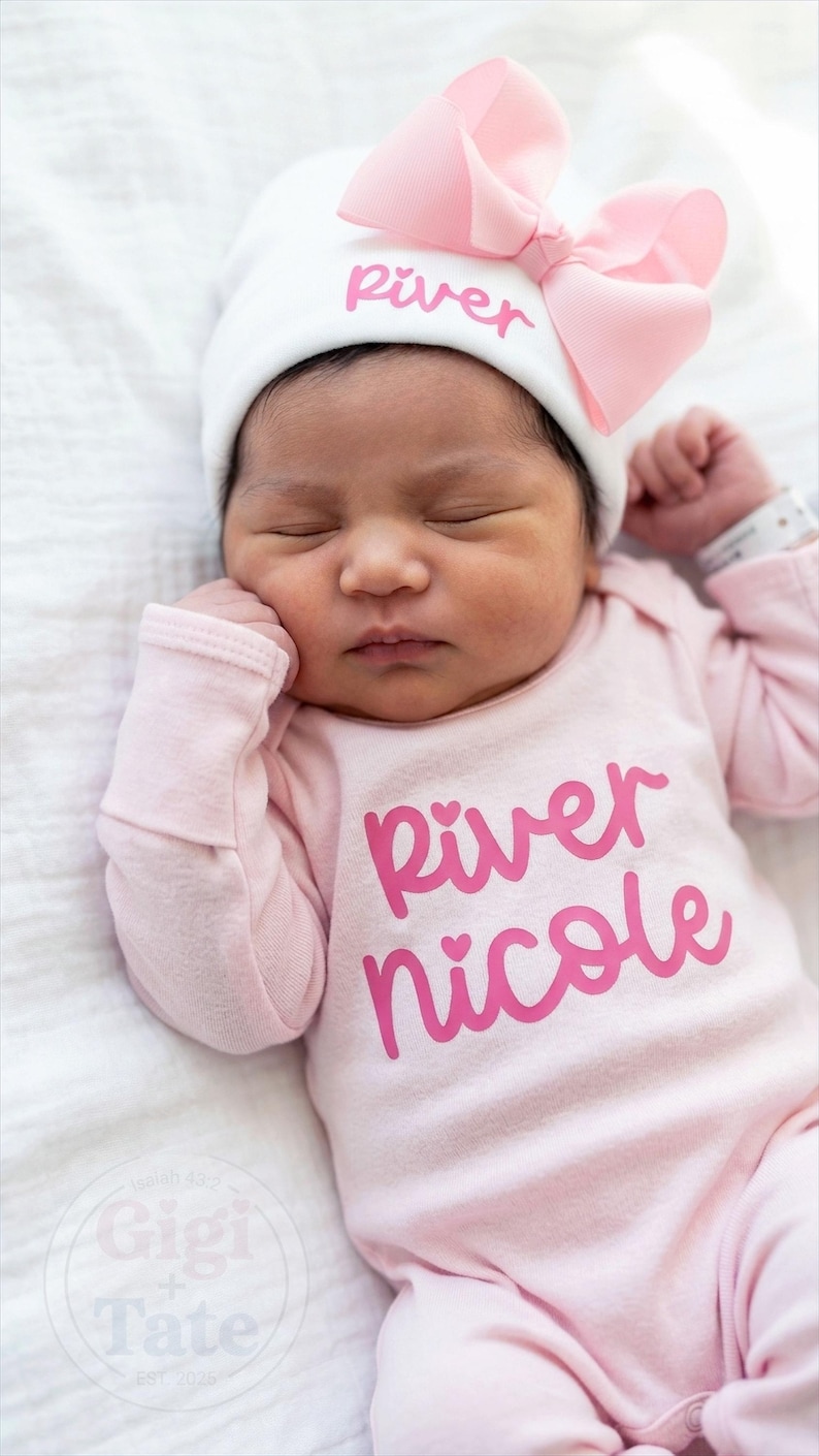 May include: A white newborn hat featuring a light pink bow and the name River embroidered in pink, paired with a long-sleeved light pink onesie printed with the name River Nicole in a bold pink script font with small heart accents over the i letters.