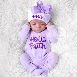 May include: A light purple long-sleeved footed onesie with the text "Holli Faith" in dark purple script. The matching white beanie features a large light purple bow and the name "Holli" in dark purple script on the front of the hat.