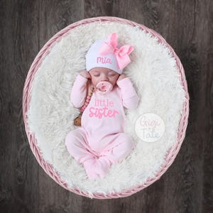 May include: A newborn baby wearing a light pink long-sleeved footed sleeper with the text little sister in pink, a white beanie with the name Mia in pink, and a large pink bow. A pink pacifier is in the baby's mouth.