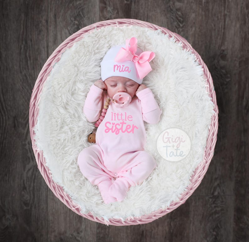 May include: A newborn baby wearing a light pink long-sleeved footed sleeper with the text little sister in pink, a white beanie with the name Mia in pink, and a large pink bow. A pink pacifier is in the baby's mouth.