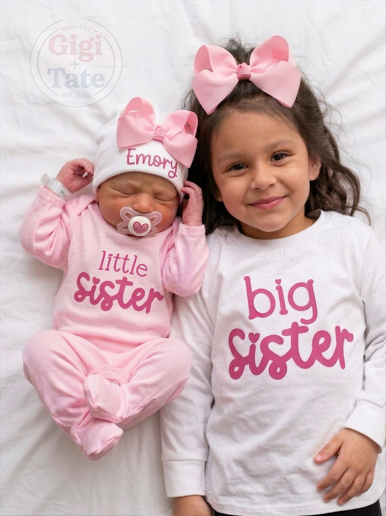 May include: A baby wearing a pink long-sleeve onesie with the text little sister and a white hat with the name Emory and a pink bow, next to a child wearing a white long-sleeve shirt with the text big sister and a pink hair bow.