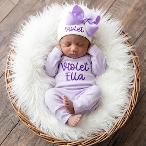 May include: A white baby hat with a large lavender bow and the name Violet in purple script, paired with a matching lavender long-sleeve onesie featuring the text Violet Ella in purple script, worn by a sleeping infant in a white faux fur basket.