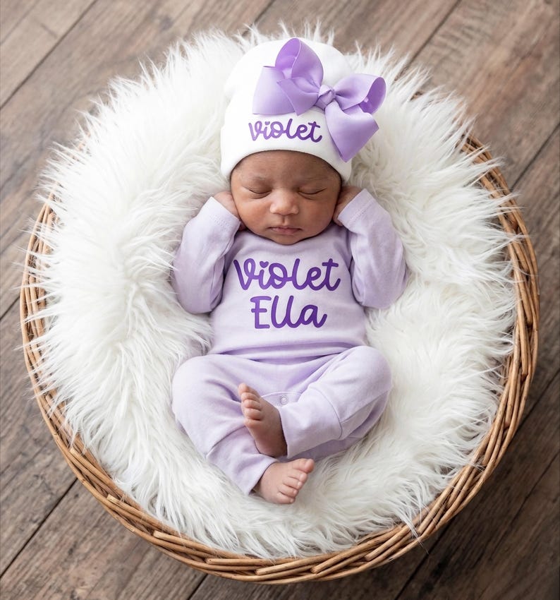 May include: A white baby hat with a large lavender bow and the name Violet in purple script, paired with a matching lavender long-sleeve onesie featuring the text Violet Ella in purple script, worn by a sleeping infant in a white faux fur basket.